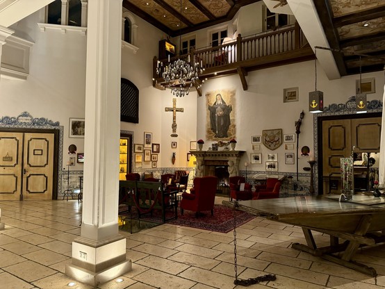 An ornately decorated room with a traditional aesthetic, featuring tiled floors, a large fireplace, assorted framed artwork on the walls, a heavy wood door, and a chandelier. The space includes a balcony level, red upholstered chairs, and religious imagery. Looks like in a monastery.
