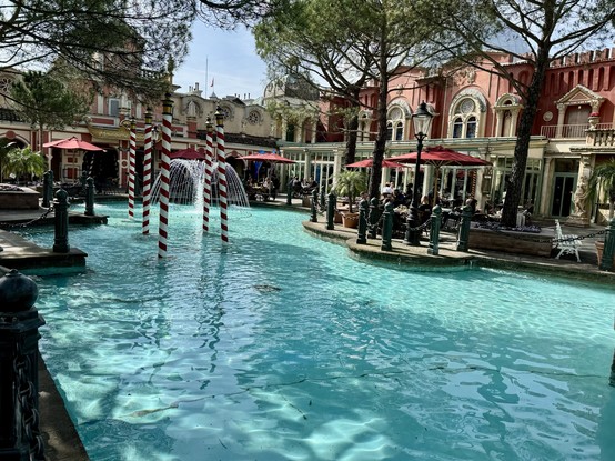 A vivid outdoor scene with a turquoise pool, red and white striped installations forming an arch over the water, and a fountain. In the background, people sit at a cafe terrace adjacent to elegant pink buildings framed by trees. Looks like in Italy.