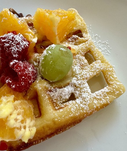 A waffle topped with fresh oranges, raspberries, a grape, and dusted with powdered sugar.