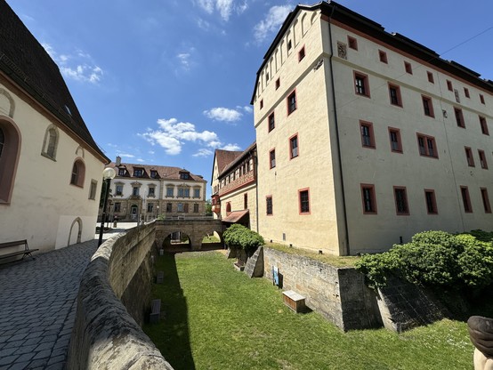 Historic European building complex with a stone wall and a pedestrian bridge over a grassy moat under a clear blue sky.