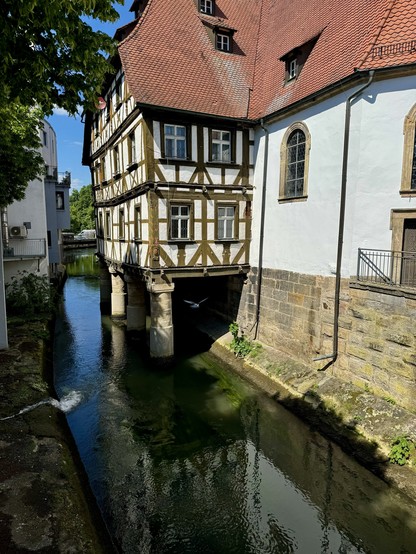 Half-timbered house extending over a narrow river, with traditional architecture, supported by stone pillars, adjacent to a larger building with a tiled roof.