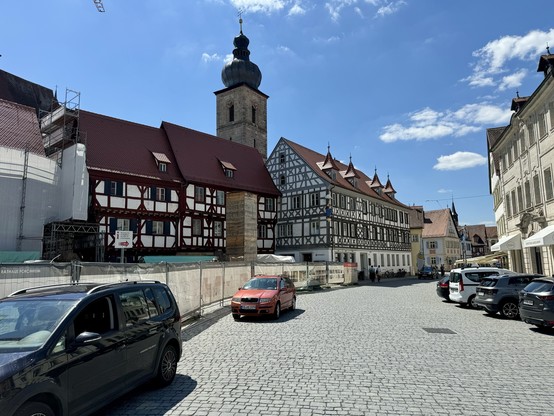A sunny day in a European town with half-timbered buildings, a cobblestone street, parked cars, and a church tower in the background. Construction scaffolding is visible on one building.