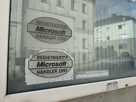 Faded and peeling stickers on a window displaying "Registrierter Microsoft Händler 1997" and "Registrierter Microsoft Händler 1998," indicating past registration as an authorized Microsoft dealer, with reflections of buildings
