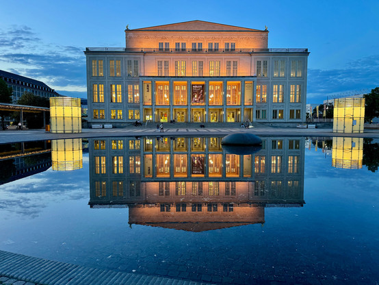 The picture shows the Leipzig Opera House in Germany. The building is illuminated at dusk, emphasising its impressive architecture. The façade of the opera house is symmetrical and has numerous large windows that are illuminated from the inside. In front of the opera house is a large, calm water area that perfectly reflects the building and enhances its majestic appearance. To the left and right of the opera house are two modern, cubic light installations, which are also illuminated and whose l…