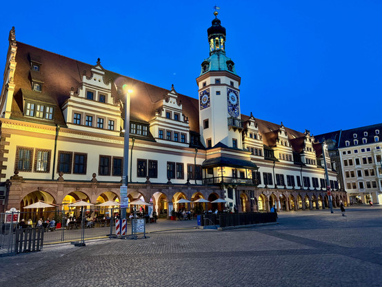 The picture shows the Old Town Hall in Leipzig, Germany. The building is a historic structure and a striking example of Renaissance architecture. It is taken in the evening hours, which is why the façade of the town hall is illuminated by warm light. The building has a steep, red roof and a façade with many windows and decorations. A tall tower with a clock and a bell tower rises in the centre of the building.

In front of the town hall are arcades under which people sit in a restaurant or café…