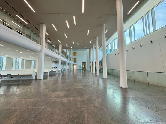 The picture shows the interior of a modern building at Leipzig University. The room is large, open and flooded with light, with high ceilings and many windows that let in plenty of daylight.

The architecture is minimalist and functional, with smooth, white walls and numerous, slender, white columns that structure and support the space. The floor consists of large, glossy tiles that give the room an elegant look.

To the left of the picture are modern, white tables and grey chairs, which probab…