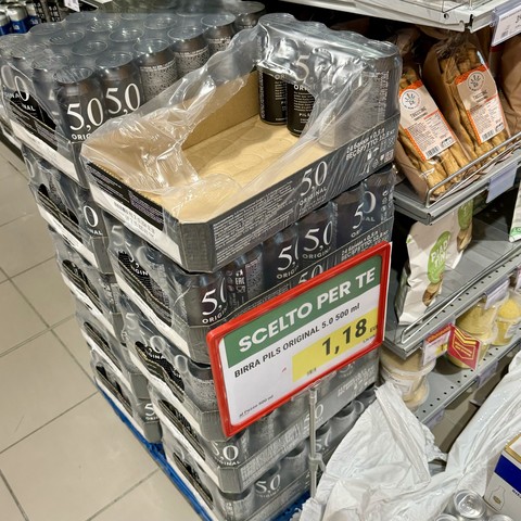 Gestapelte Dosen von 5.0 Original Beer (Birra Pils) in einem Supermarkt, mit einem Schild mit der Aufschrift "SCELTO PER TE" (für Sie ausgewählt) und einem Preis von 1,18 EUR. In der Nähe von Regalen befinden sich verschiedene