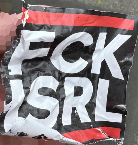 A crumpled sticker, because removed, with the inscription "FCK ISRL", held by a hand. The letters are in white on a black background, with a red stripe at the top and bottom edge.