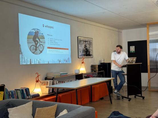 In the same room, another person is now standing at the podium, giving a presentation. On the wall, a slide titled "$ whoami" is displayed. It features a circular photo of a person on a bicycle, along with text containing personal information: Philip Laine is of Swedish-Finnish descent, lives in Berlin, works as a developer, is the founder of the "ReFlektion" project, and was previously a maintainer of the "FLUX" tool. It’s also humorously noted that he was once nearly recruited by Microsoft.

…