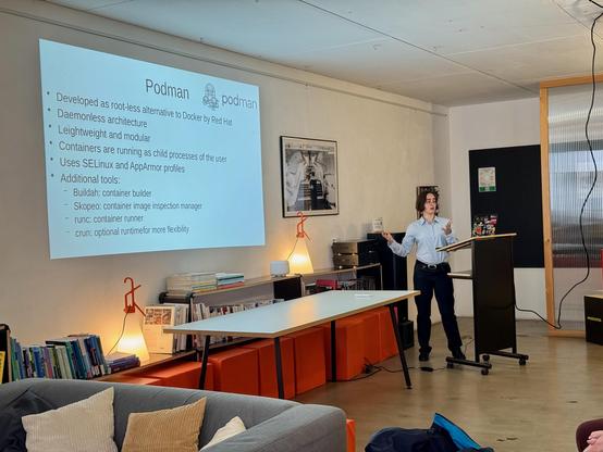 The scene depicts a bright room with cozy, modern furnishings. A person stands on the right side of the frame behind a simple podium, delivering a presentation. A slide with the title "Podman" is projected on the wall. It states that Podman is a rootless alternative to Docker, developed by Red Hat. Additional points cover the architecture, security through SELinux and AppArmor, as well as supplementary tools like Buildah, Skopeo, runc, and crun.

On the left side of the room, there is a gray so…