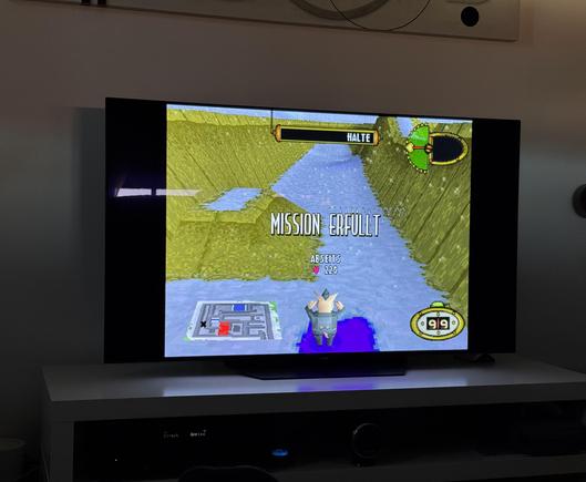 A screen in a living room displays the PlayStation game "Frontschweine" with the message "MISSION COMPLETE." A virtual character is visible in the game, moving through a landscape. Various displays and menus are shown on the screen. The TV sits on a white piece of furniture.