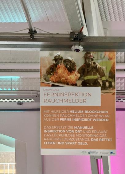 Ein Poster hängt an einer Decke. Das obere Bild zeigt Feuerwehrleute in Schutzkleidung mit Flammen im Hintergrund. Der Text auf dem Poster beschreibt eine Lösung zur Ferninspektion von Rauchmeldern mithilfe der Helium-Blockchain. Diese Methode erlaubt die Inspektion ohne WLAN und ersetzt die manuelle vor Ort Inspektion. Dies ermöglicht lückenloses Monitoring, rettet Leben und spart Geld.