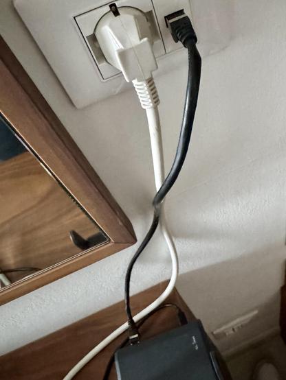 A white power cable and a black Ethernet cable are plugged into a wall socket. A small mirror can be seen next to the socket and the cables lead to a wifi access point on a piece of wooden furniture.