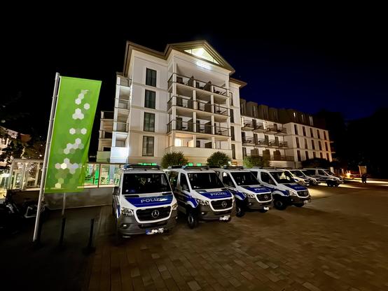 Ein modernes Hotelgebäude bei Nacht mit einem großen grünen Banner links im Bild. Vor dem Gebäude stehen mehrere geparkte Polizeifahrzeuge in einer Reihe.