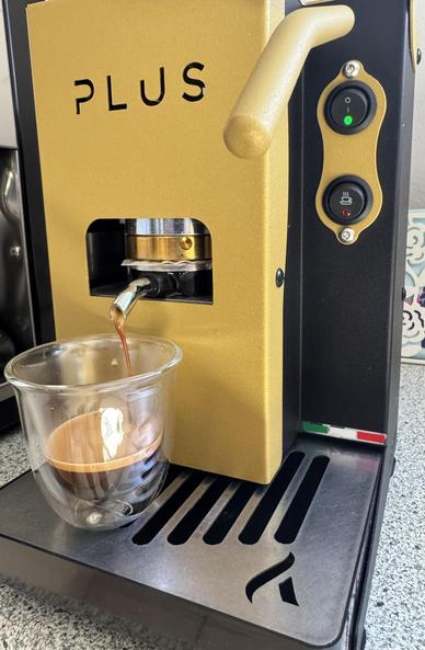 Eine goldene Espressomaschine mit der Aufschrift "PLUS" gießt Espresso in ein doppelwandiges Glas. Auf der rechten Seite sind zwei Schalter mit Anzeigen zu sehen, und auf dem unteren Rost befindet sich eine italienische Flagge.