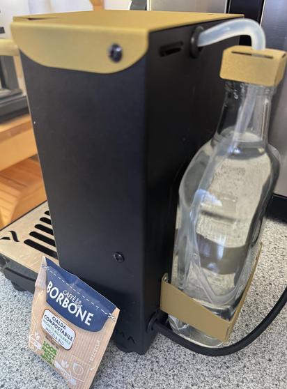 Eine Maschine auf einer Küchenarbeitsplatte mit einer Glasflasche, die an der Seite befestigt ist. Ein kleiner Schlauch verbindet die Flasche mit der Maschine. Vor der Maschine liegt ein Päckchen Kaffee mit der Aufschrift "BORBONE."