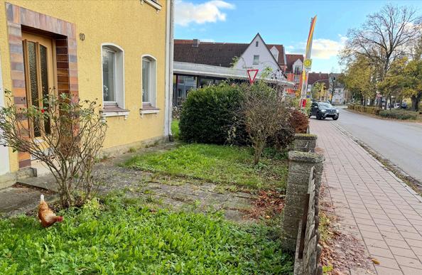 The picture shows the front yard of a yellow house on a quiet, small street. In the left foreground, a brown and white chicken is visible on a patch of green, seemingly looking for food. To its left, a bare rose bush stands directly in front of the house entrance, which has a wooden door with a colorful frame.

In the right part of the image, there is a low stone fence with more shrubs and bushes behind it. A paved sidewalk runs behind the yard, parallel to a street with little traffic. A black…