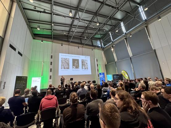 A presentation in a modern conference hall: A person is speaking at a lectern in front of a packed audience, with a large screen behind them showing a slide titled "OSS Virtualization & IaaS" along with a schematic diagram of open-source cloud infrastructure. To the right and left are information boards and exhibition booths with green light accents, and several empty chairs are waiting on the stage for a panel discussion. The high, bright room with metal beams, hanging spotlights, and slender …
