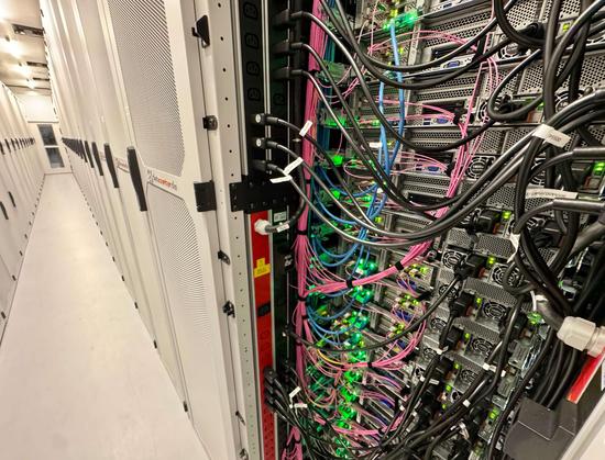 The picture shows a long, brightly lit hallway in a data center. On the right, several server racks are open, showing lots of servers packed tightly together with tons of cable connections. The cables are different colors—lots of black ones, some blue, and some pink. They're all neatly bundled and connect the backs of the servers. Green indicator lights are glowing on the servers, which means they're up and running. The left side of the picture shows closed rack doors with ventilation grilles a…