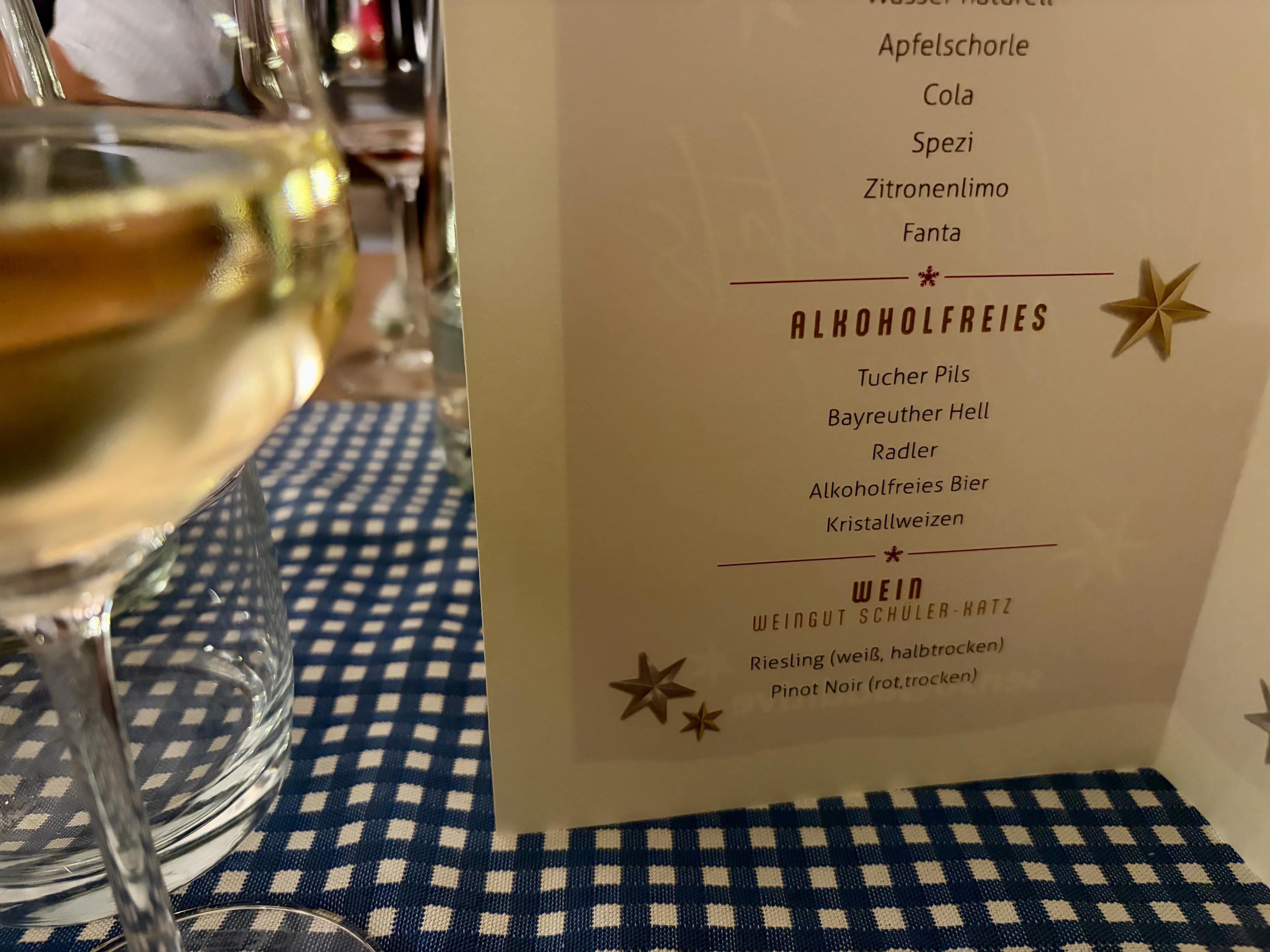 Auf dem Foto steht auf einem blau-weiß karierten Tisch eine Weinglas mit weißem Wein im Vordergrund links. Rechts daneben steht eine aufklappbare Getränkekarte aus cremefarbenem Karton, auf der in dunkelroter Schrift verschiedene Getränke aufgelistet sind.
Im oberen Bereich der sichtbaren Seite der Karte stehen alkoholfreie Getränke wie Wasser, Apfelschorle, Cola, Spezi, Zitronenlimo und Fanta. Darunter, durch eine Linie abgesetzt, folgt die Überschrift „Alkoholfreies“ mit Biersorten wie Tucher Pils, Bayreuther Hell, Radler, alkoholfreies Bier und Kristallweizen. Ganz unten steht die Rubrik „Wein“ mit dem Hinweis auf das Weingut Schuler-Katz sowie zwei Weine: ein halbtrockener weißer Riesling und ein trockener roter Pinot Noir. Im Hintergrund sind weitere Gläser und verschwommene Personen angedeutet, was auf eine Restaurant- oder Feieratmosphäre schließen lässt.