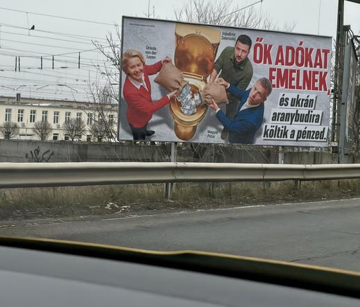 This is a large political poster on a street. On it, three well-known people are photoshopped: on the left Ursula von der Leyen, in the middle Volodymyr Zelenskyy, and on the right Péter Magyar. All three are tipping bags of money into an open golden toilet bowl, which already contains many banknotes.
The Hungarian text reads approximately:
"Ők adókat emelnek, és ukrán aranybudira költik a pénzed."
Literally translated: "They raise taxes and spend your money on a Ukrainian gold toilet."
The mes…