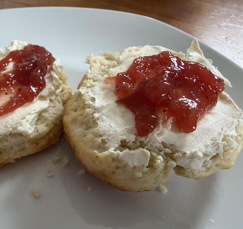 Two halves of a freshly baked scone sit on a white plate. Each scone half is generously covered with smooth white cream and topped with a glossy spoonful of red strawberry jam, and one half has a small bite taken out of it.