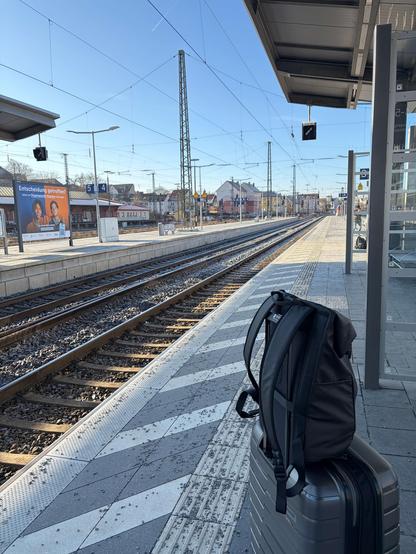 The image shows a quiet train station platform on a clear, sunny day. Two railway tracks run straight into the distance, creating strong perspective lines. Overhead, there are many electric cables supported by tall metal poles. The sky is bright blue with no clouds.

On the right side of the platform, close to the camera, there is a medium-sized gray hard-shell suitcase. A black backpack is attached on top of it, standing upright near the edge of the platform. The platform surface has light and…