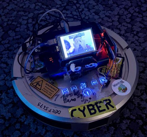 The image shows a circular robotic vacuum cleaner placed on a dark, patterned carpet. The robot has been heavily modified and looks experimental. On top of it sits a small rectangular screen displaying a paused video with two people; one person is covering their face with their hands. Several cables and electronic components are connected to the screen and spread across the top of the robot.

The surface of the vacuum cleaner is covered with stickers, labels, and handwritten markings. Visible s…