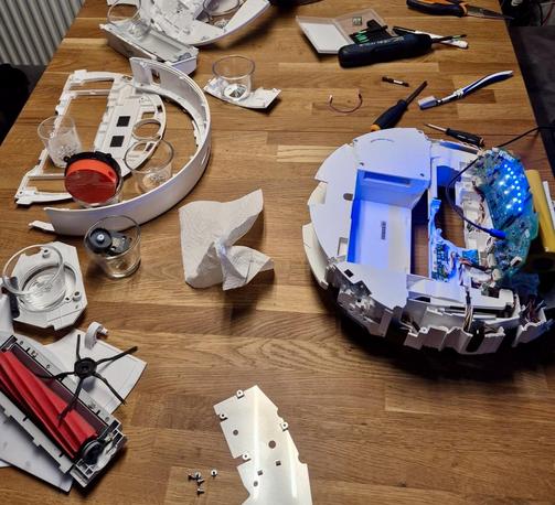 A partially disassembled robot vacuum is spread out on a wooden table. Various internal components are visible, including circuit boards with glowing blue lights, a red and black brush attachment, and other plastic parts. Several small glasses, tools like screwdrivers, and a paper towel are also on the table, indicating a repair or maintenance process in progress.