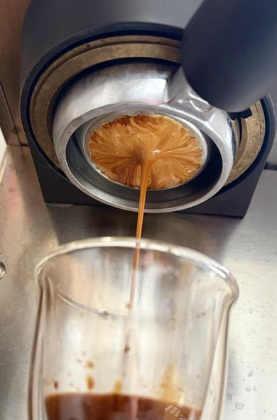 Close-up of espresso being brewed. Coffee is flowing from the portafilter into a clear glass cup, with visible layers forming in the espresso shot. The machine and cup are on a metal surface.