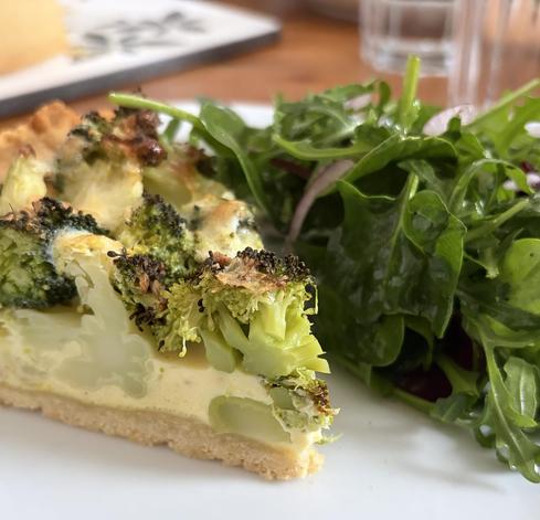 A slice of broccoli quiche with a golden crust is on a white plate next to a fresh green salad. The salad includes arugula and thin slices of red onion. The plate rests on a wooden table, and a glass is visible in the background.