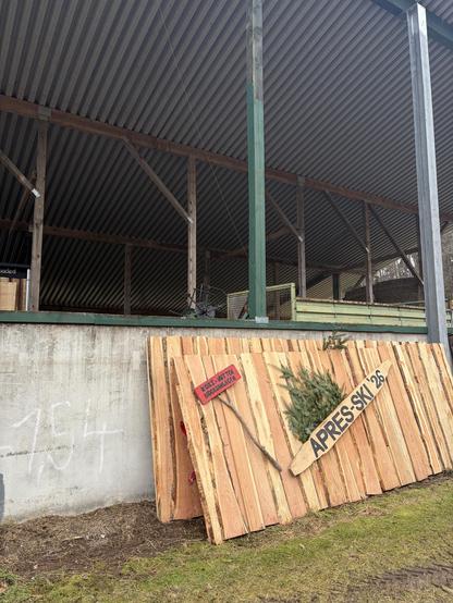 Blick von der Seite Ein rustikaler hölzerner Hintergrund steht vor einer Betonwand und zeigt zwei Schilder. Ein Schild besteht aus einem Stück Holz in Form eines Skis, auf dem in fetten Buchstaben „APRES-SKI ’26“ steht. Das andere Schild ist kleiner, rot bemalt und an einem Ast befestigt, mit der Aufschrift „KOKS-NUTTEN BIOGASANLAGEN“. Hinter dem Skischild sind Tannenzweige gesteckt, und im Vordergrund befindet sich ein Stück Rasen. Hintergrund: Landwirtschaftliche Geräte